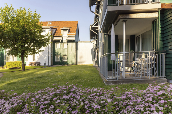 Garten mit Blumenteppich vor einem Gebäude mit Balkon bei sonnigem Wetter.