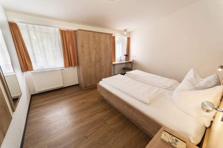 Schlafzimmer mit Doppelbett, Schrank, Fenster mit Vorhängen und Holzboden.