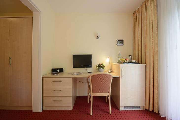 Schreibtisch mit Computer, Lampe, Stuhl und Minibar in einem Hotelzimmer.