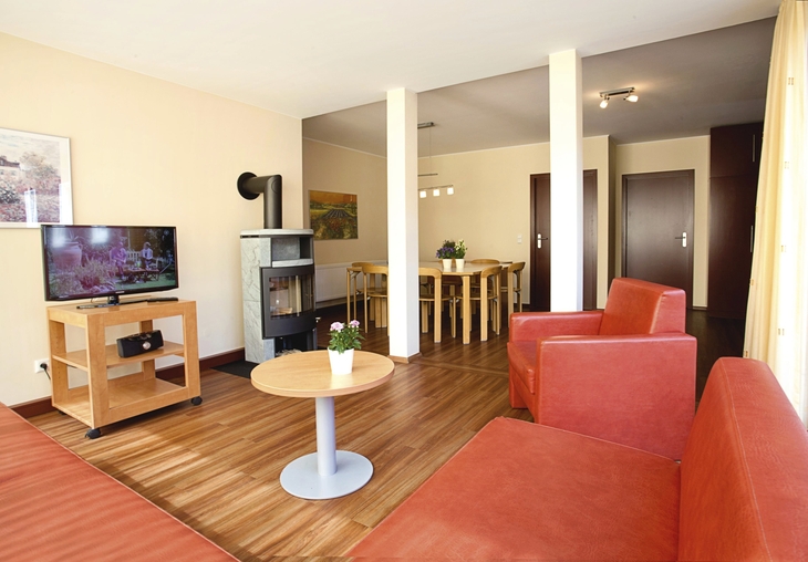 Wohnzimmer mit rotem Sofa, Ofen, Fernseher und Esstisch in der Ferienanlage Prüm