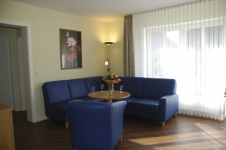 Wohnbereich mit blauer Couch, Sessel, rundem Tisch und großem Fenster.