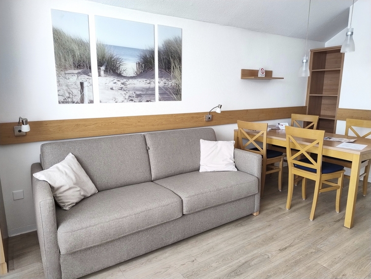 Wohnzimmer mit grauem Sofa, Esstisch und Stühlen, Wandbild mit Strandmotiv.