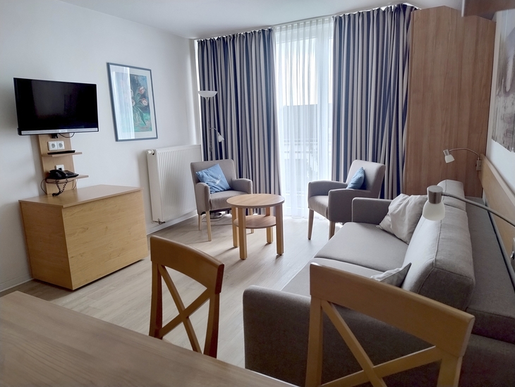 Modernes Wohnzimmer mit Sofa, Sessel, Tisch und Fernseher vor großen Vorhängen.