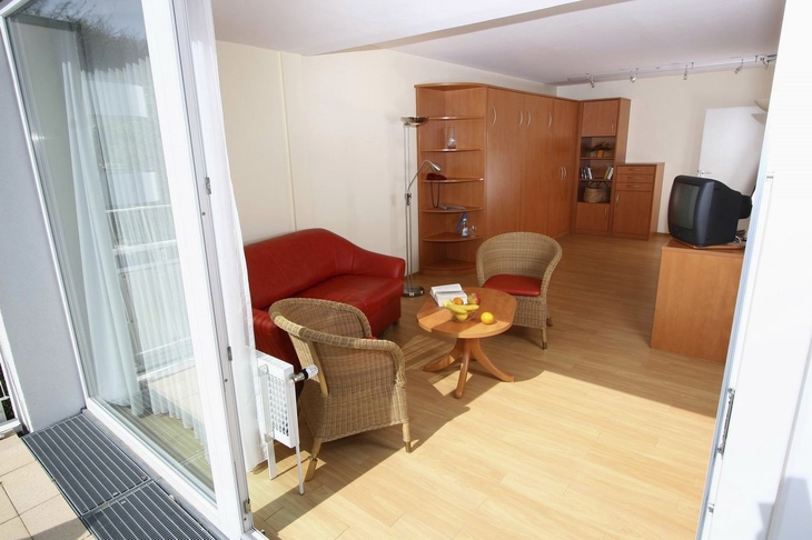 Ein Wohnzimmer mit Sofa, zwei Sesseln, einem Tisch, Fernseher und offener Balkontür.