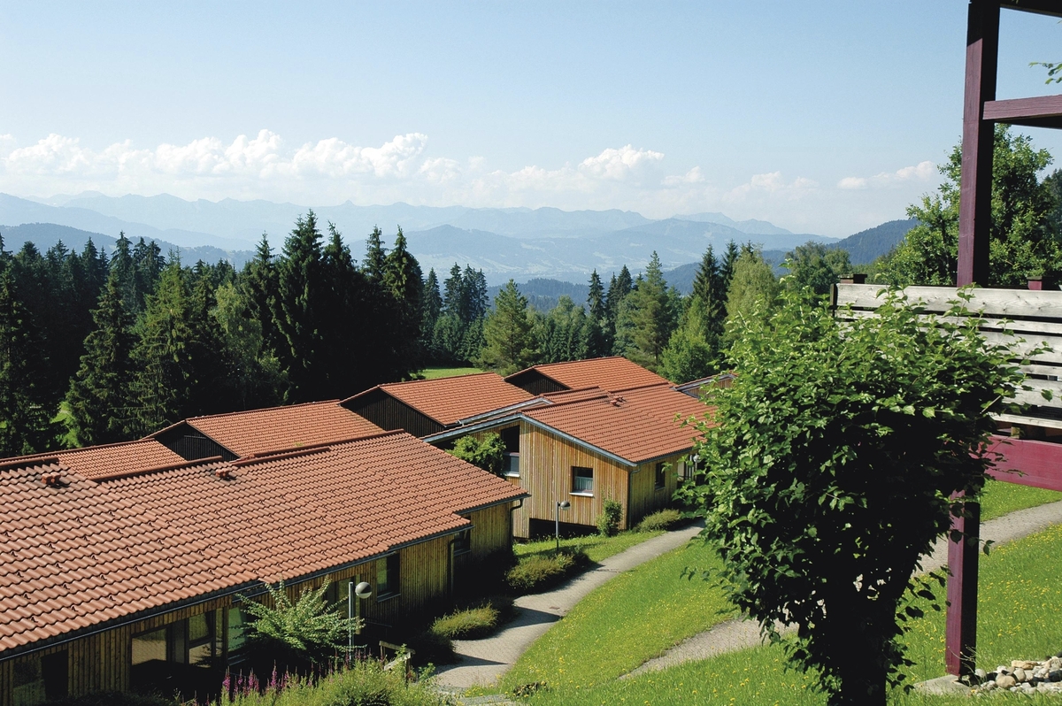 Häuser mit roten Dächern vor Alpenpanorama und bewaldeter Landschaft.