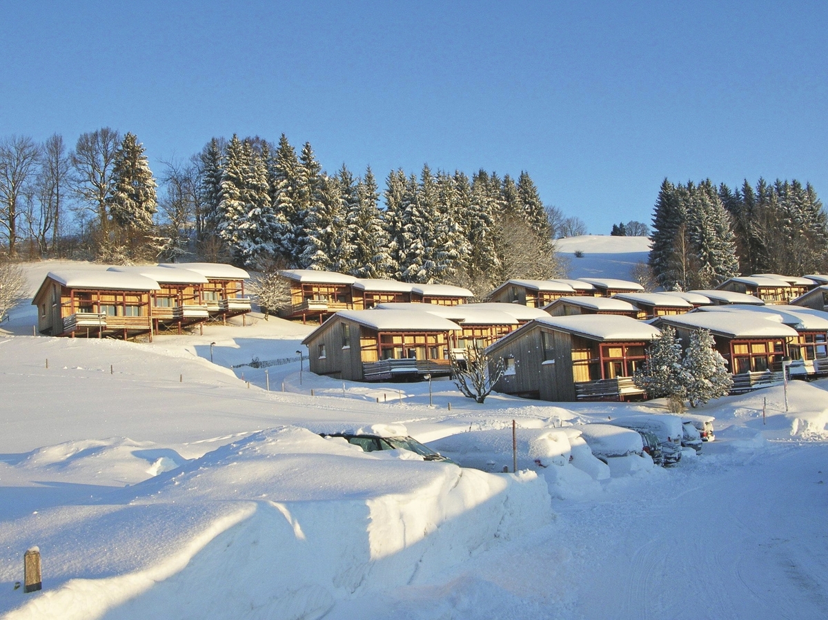 Verschneite Hüttenlandschaft in Scheidegg mit schneebedeckten Bäumen bei klarem Himmel.