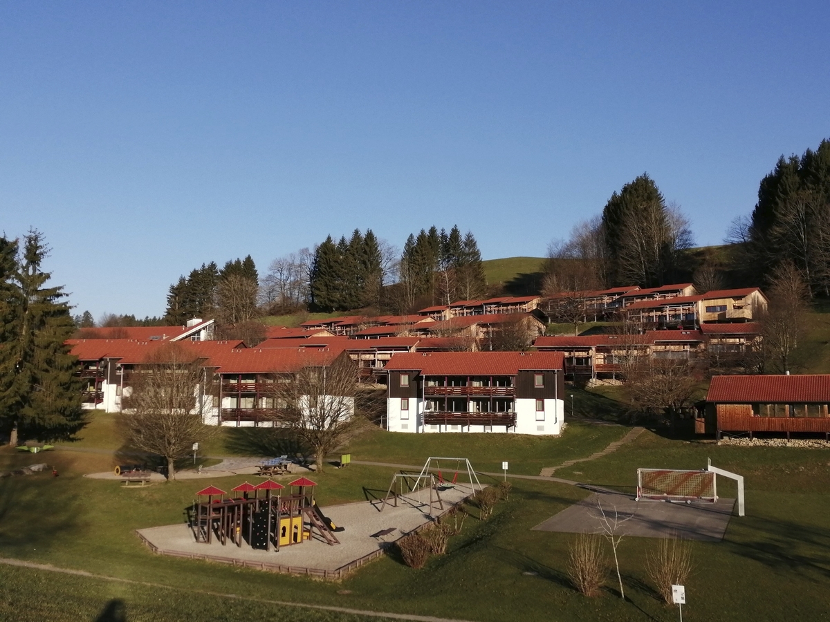 Blick auf einen Spielplatz vor Wohnhäusern mit rotem Dach in einer grünen Landschaft.