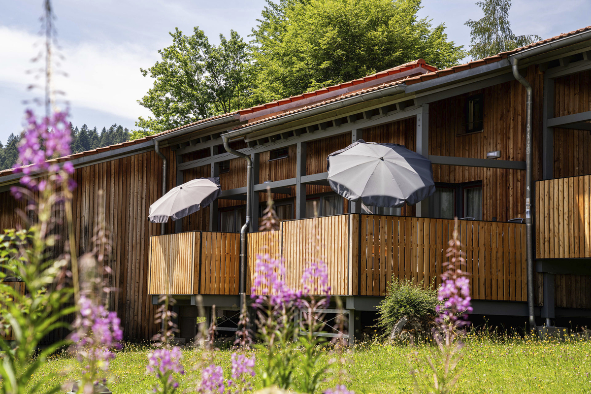 Holzgebäude mit Balkonen und Sonnenschirmen, umgeben von blühenden Pflanzen.