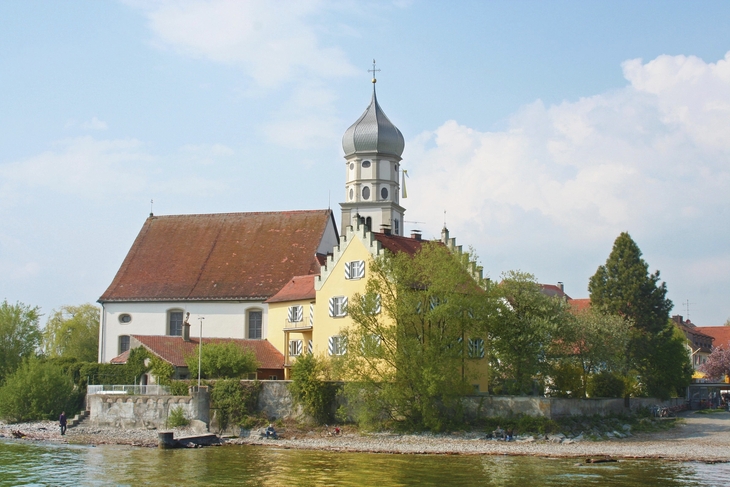 Wasserburg mit Kirchturm und Bäumen am Ufer eines Sees unter blauem Himmel mit weißen Wolken.