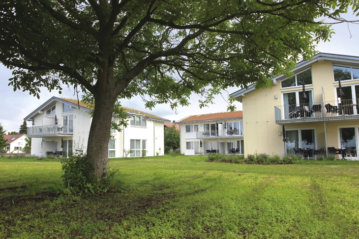 Drei Appartementhäuser an der Obstwiese, der Ferienanlage Lindau, mit Balkonen und Terrassen mit Outdoormöbeln ausgestattet und einem Baum auf einer grünen Wiese im Vordergrund.