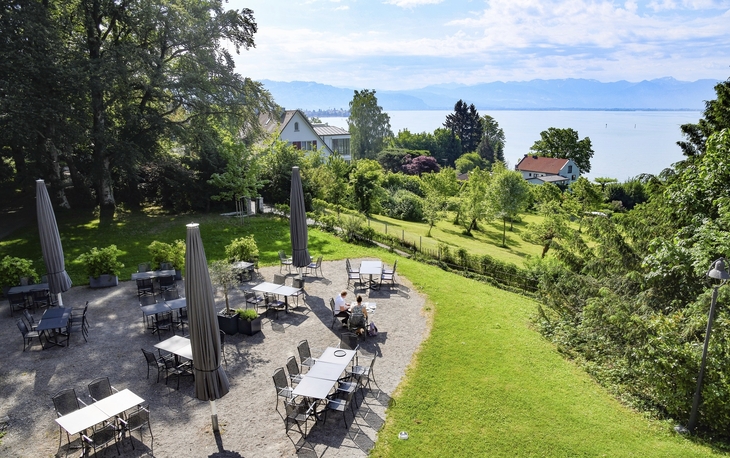 Biergarten der Villa Alwind, in der Ferienanlage Lindau, mit Tischen und Stühlen, sowie geschlossenen Sonnenschirmen, umgeben von Bäumen und einer Wiese mit Blick auf den Bodensee. bei leichtbewölktem Himmel.