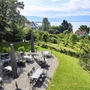 Biergarten der Villa Alwind, in der Ferienanlage Lindau, mit Tischen und Stühlen, sowie geschlossenen Sonnenschirmen, umgeben von Bäumen und einer Wiese mit Blick auf den Bodensee. bei leichtbewölktem Himmel.