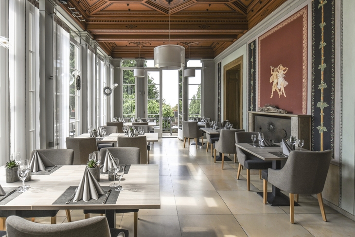 Elegantes Restaurant in der Villa Alwind, in der Ferienanlage Lindau, mit eingedeckten Tischen, grauen gepolsterten Stühlen und großen weißen Fenstern, sowie kunstvoller Holzdecke.