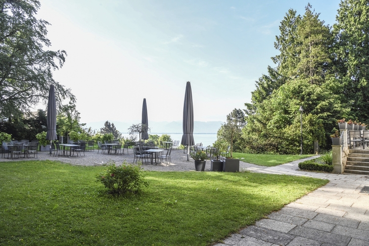 Biergarten der Villa Alwind, in der Ferienanlage Lindau, mit Tischen und Stühlen, sowie geschlossenen Sonnenschirmen, umgeben von Bäumen und einer Wiese mit Blick auf den Bodensee. bei leicht bewölktem Himmel.