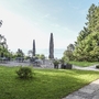 Biergarten der Villa Alwind, in der Ferienanlage Lindau, mit Tischen und Stühlen, sowie geschlossenen Sonnenschirmen, umgeben von Bäumen und einer Wiese mit Blick auf den Bodensee. bei leicht bewölktem Himmel.