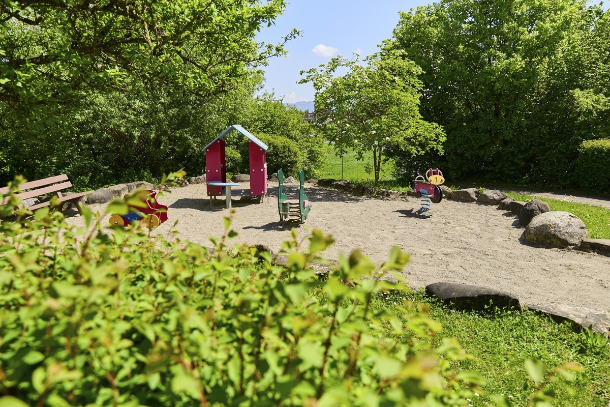 Kleiner Spielplatz mit Wipptieren und Häuschen sowie einer Parkbank umgeben von Bäumen bei sonnigem Wetter in der Ferienanlage Lindau.