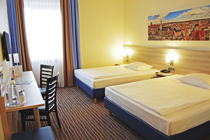 Hotelzimmer mit zwei Einzelbetten, Schreibtisch und großem Stadtbild an der Wand.