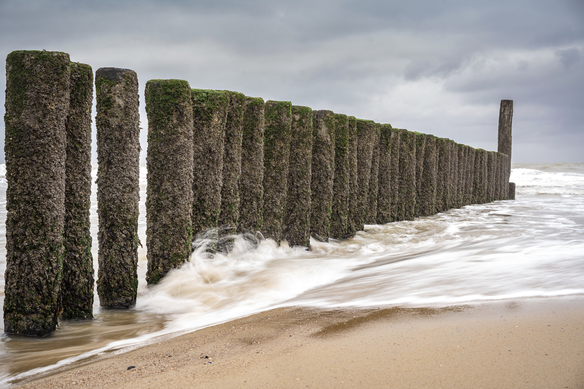 Wellen treffen auf eine Reihe hölzerner Buhnen am Strand bei bewölktem Himmel.