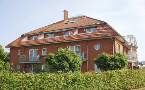 Rotes Backsteingebäude mit Balkon und rotem Dach, umgeben von grüner Hecke.
