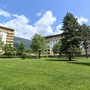 Appartementanlage mit grüner Wiese und Bäumen vor Bergen unter blauem Himmel.