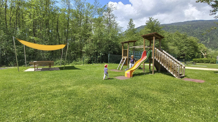 Spielplatz mit Rutsche und Kindern auf Rasenfläche vor bewaldeter Bergkulisse.