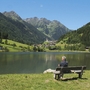 Eine Person sitzt auf einer Bank am See, umgeben von Bergen.