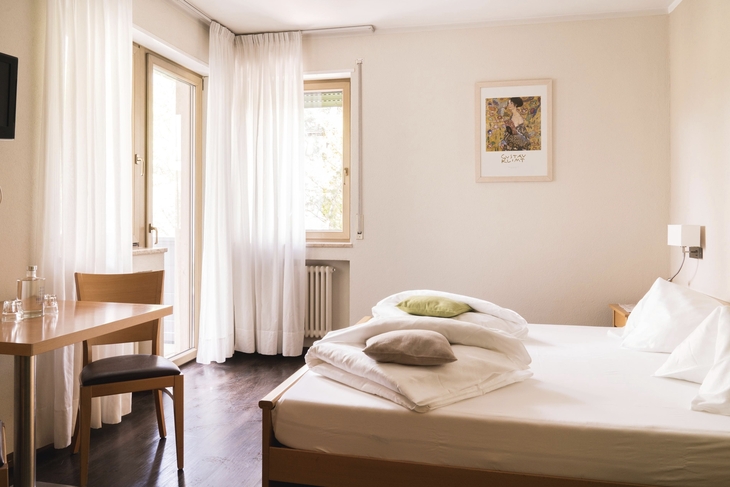 Doppelzimmer mit Bett, Tisch und Stuhl, helle Vorhänge und Bild an der Wand.