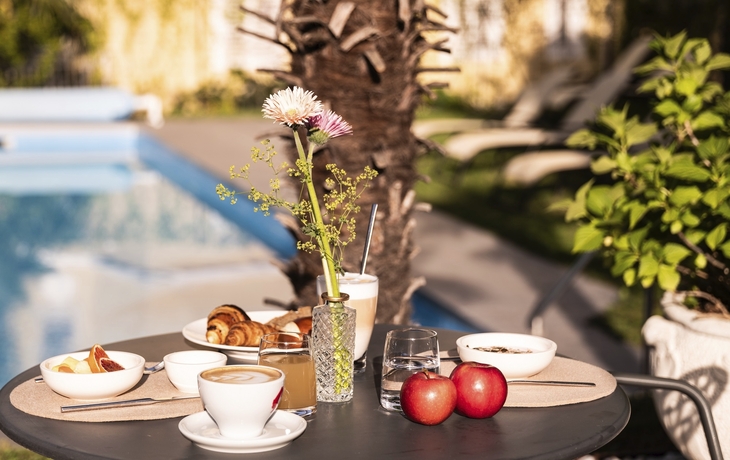 Frühstückstisch auf Terrasse mit Getränken und Gebäck, Blick auf Pool und Pflanzen.