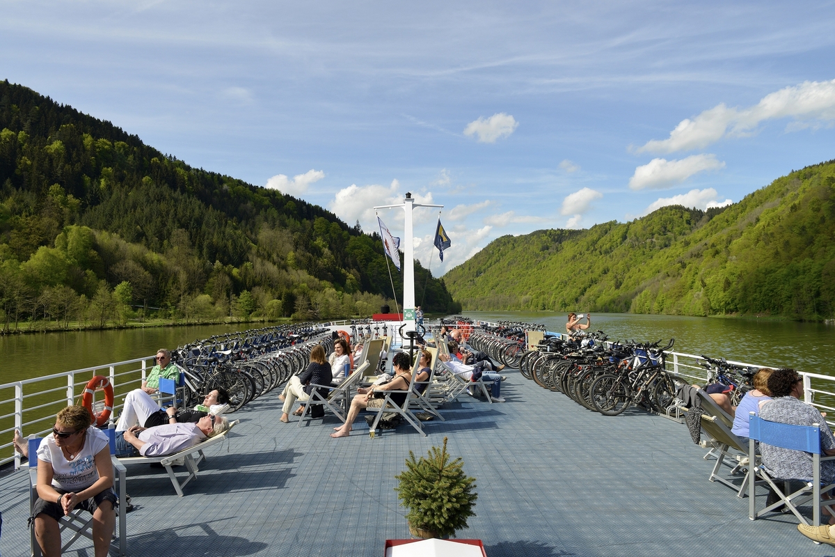 Passagiere entspannen auf dem Sonnendeck eines Flussschiffs, umgeben von grünen Hügeln.