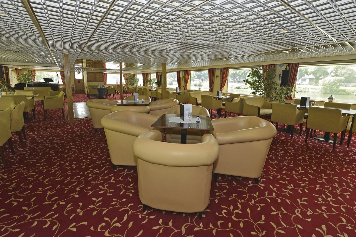 Schicker Salon mit Sesseln und Tischen auf einem Flussschiff, rote Teppiche und große Fenster.