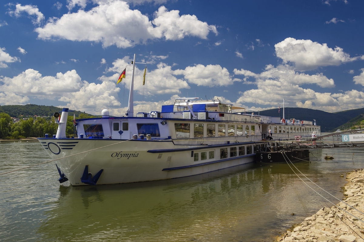 Flusskreuzfahrtschiff MS Olympia am Ufer mit blauem Himmel und Wolken im Hintergrund.