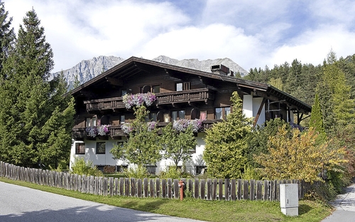 Landhaus mit Holzbalkon und Blumen, umgeben von Bäumen und Berglandschaft im Hintergrund.