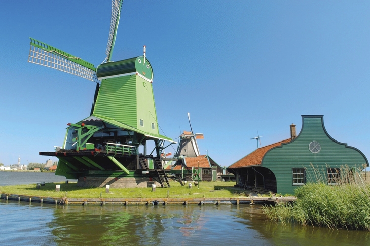 Grüne Windmühle an einem Flussufer bei sonnigem Wetter.
