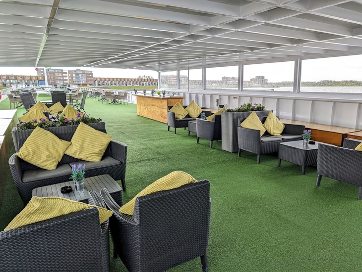 Sonnendeck auf einem Schiff mit Sitzgruppen und Blick auf Gebäude in der Ferne.