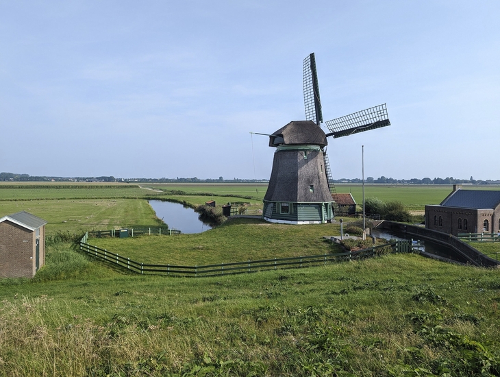 Windmühle in nordholländischer Landschaft mit Kanal und Gebäuden im Hintergrund.