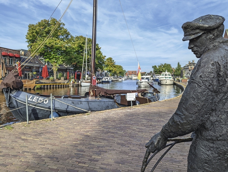 Statue eines Mannes am Kai, Boote auf Wasserweg, Bäume und Gebäude im Hintergrund