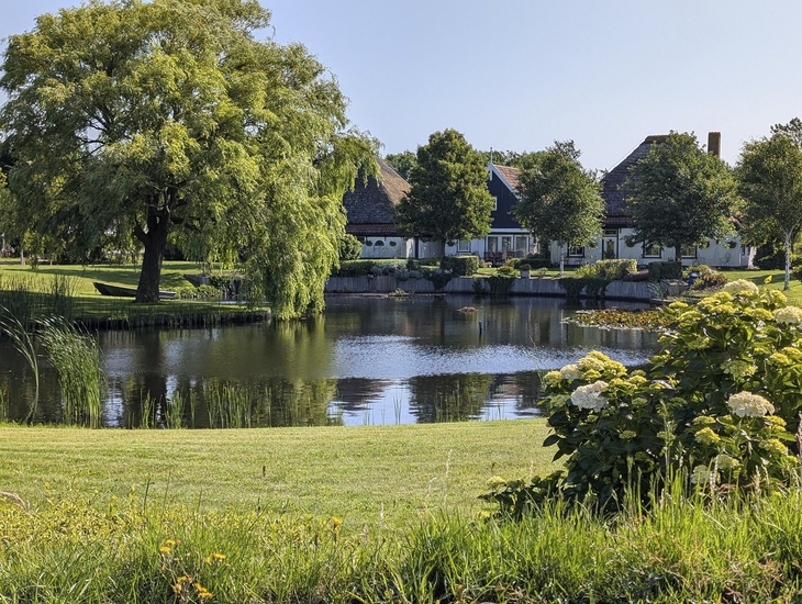 Grüne Landschaft mit Teich, Bäumen und Häusern am Ufer unter blauem Himmel.