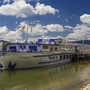 Flusskreuzfahrtschiff MS Olympia am Ufer mit blauem Himmel und Wolken im Hintergrund.