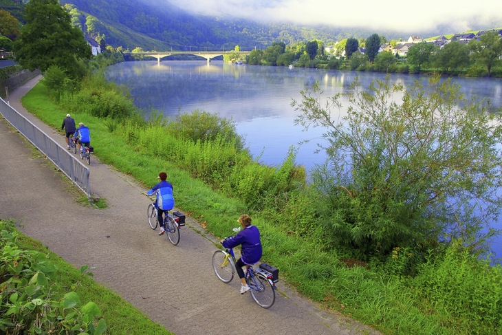 Radfahrer auf einem Weg entlang eines Flusses mit Brücke im Hintergrund.