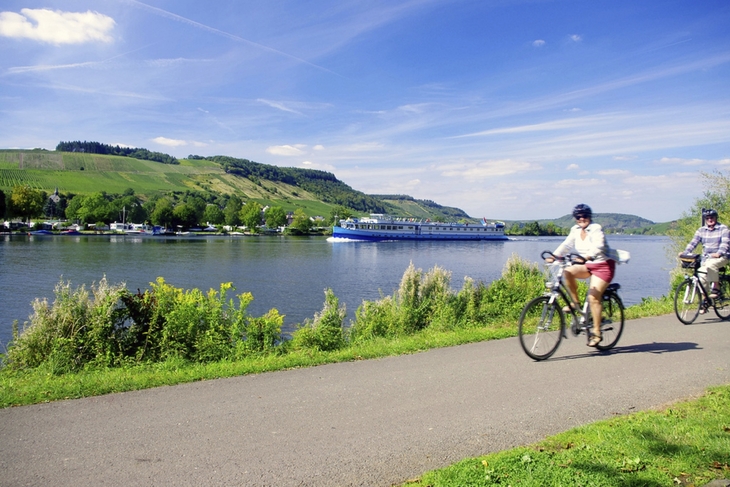 Radfahrer auf einem Weg am Fluss Mosel vor einem Schiff, Landschaft im Hintergrund.