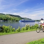Radfahrer auf einem Weg am Fluss Mosel vor einem Schiff, Landschaft im Hintergrund.