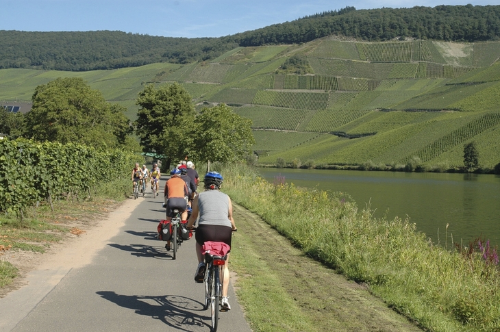Radfahrer auf einem Weg entlang eines Flusses in einer grünen, hügeligen Landschaft.