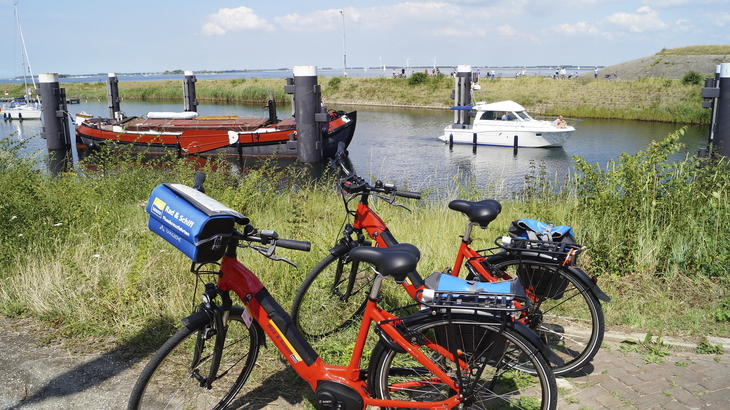 Rote Fahrräder am Ufer mit Booten im Hintergrund vor sonniger Küstenlandschaft.