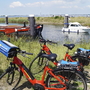 Rote Fahrräder am Ufer mit Booten im Hintergrund vor sonniger Küstenlandschaft.
