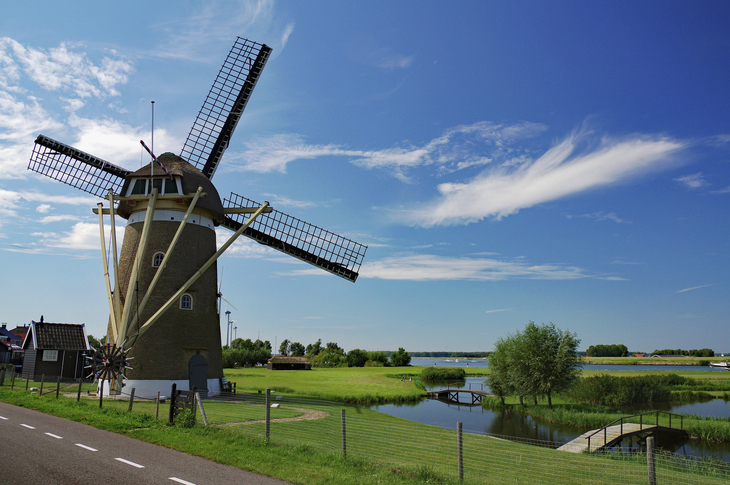 Windmühle am Flussufer unter blauem Himmel und grüner Landschaft.