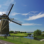 Windmühle am Flussufer unter blauem Himmel und grüner Landschaft.