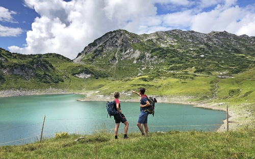 Zwei Wanderer am Formarinsee vor einer Bergkulisse bei sonnigem Wetter.