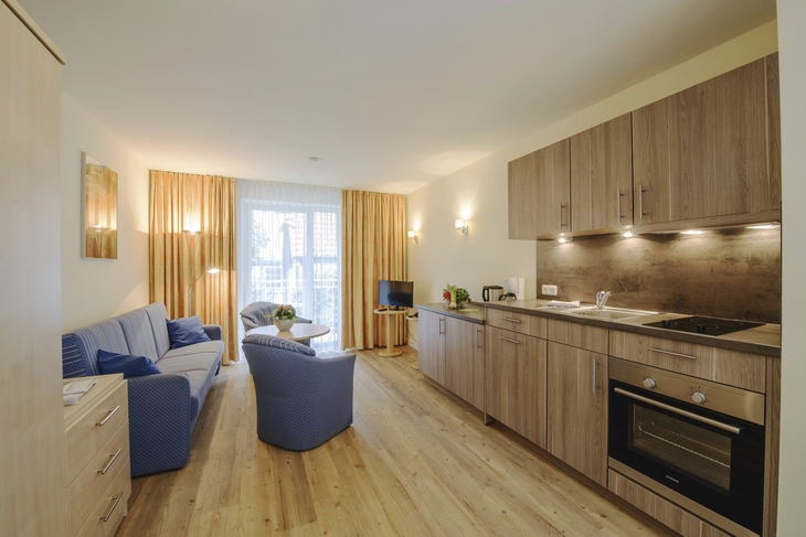 Wohnbeispiel Haus Oldenburg in der Ferienanlage Timmendorfer Strand. Rechts im Bild eine moderne Küchenzeile, links blaues Sofa und zwei Sessel mit kleinem runden Tisch. Hinten im Bild ein großes Fenster mit Vorhängen rechts und links und einem Fernseher davor.
