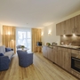 Wohnbeispiel Haus Oldenburg in der Ferienanlage Timmendorfer Strand. Rechts im Bild eine moderne Küchenzeile, links blaues Sofa und zwei Sessel mit kleinem runden Tisch. Hinten im Bild ein großes Fenster mit Vorhängen rechts und links und einem Fernseher davor.