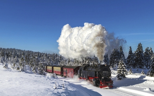 Dampflok fährt durch verschneite Landschaft mit Nadelbäumen unter klarem, blauem Himmel.